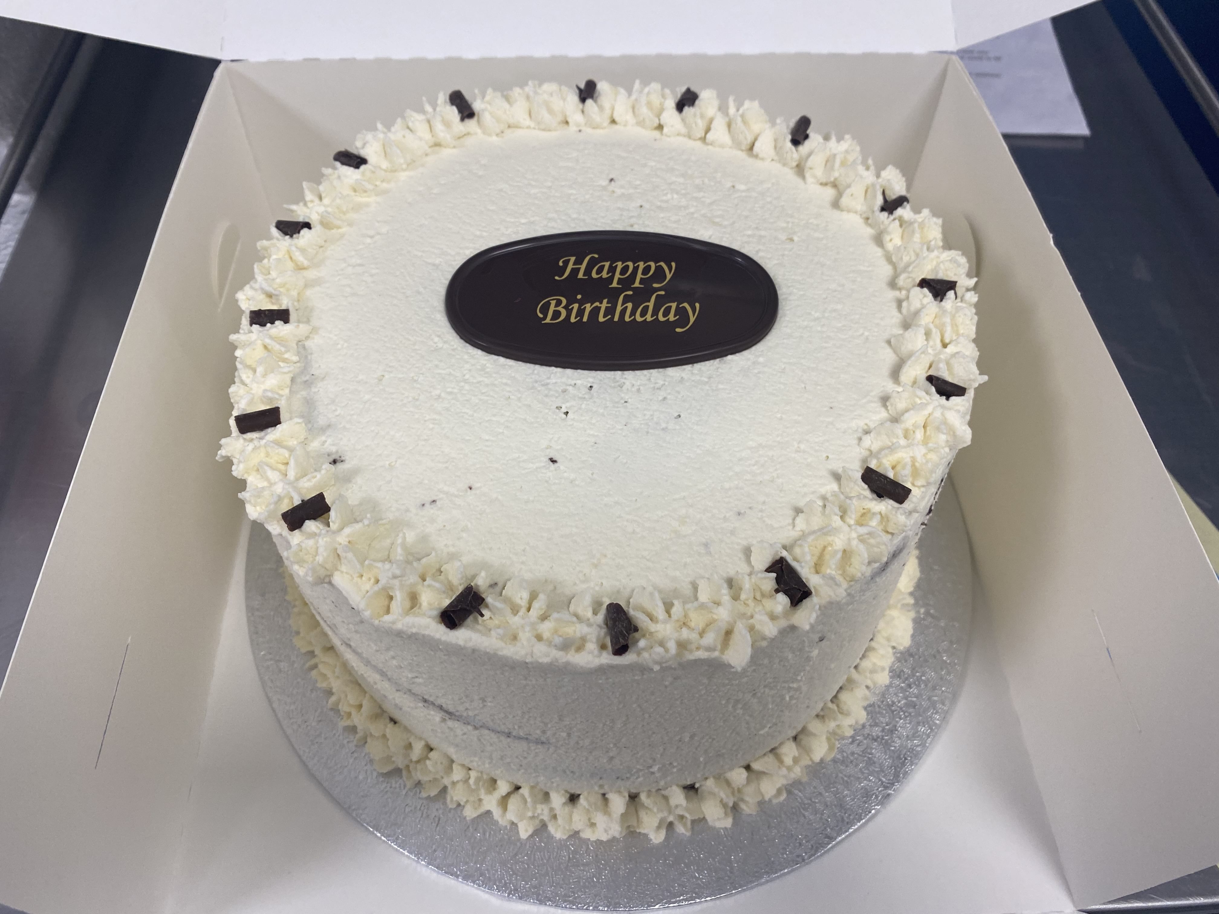 A round white frosted cake with an oval chocolate topper in the center that reads 'Happy Birthday' in gold text. The cake, reminiscent of a Black Forest cake, is decorated with piped white frosting along the top edge and small chocolate pieces. It is placed in a white cake box.